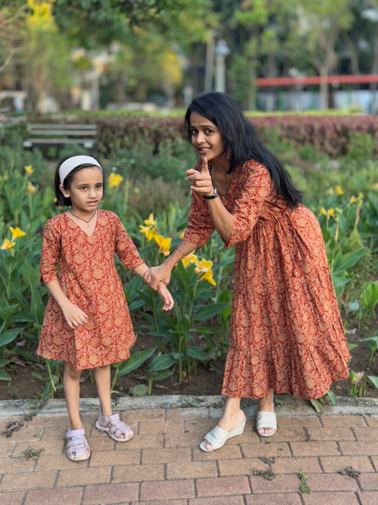 Mother Daughter in matching Twinning Light rust Colour dresses standing in a park with greenery and buildings in the background.